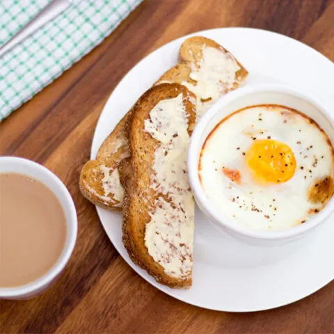 A small white bowl of baked eggs, ham and tomatoes on a white plate with toast and butter. Set on a deep wooden backdrop with a cup of tea, a green checked tea towel and a spoon.