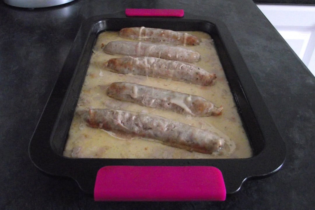 sausages lined up in a roasting tray covered in yorkshire pudding batter