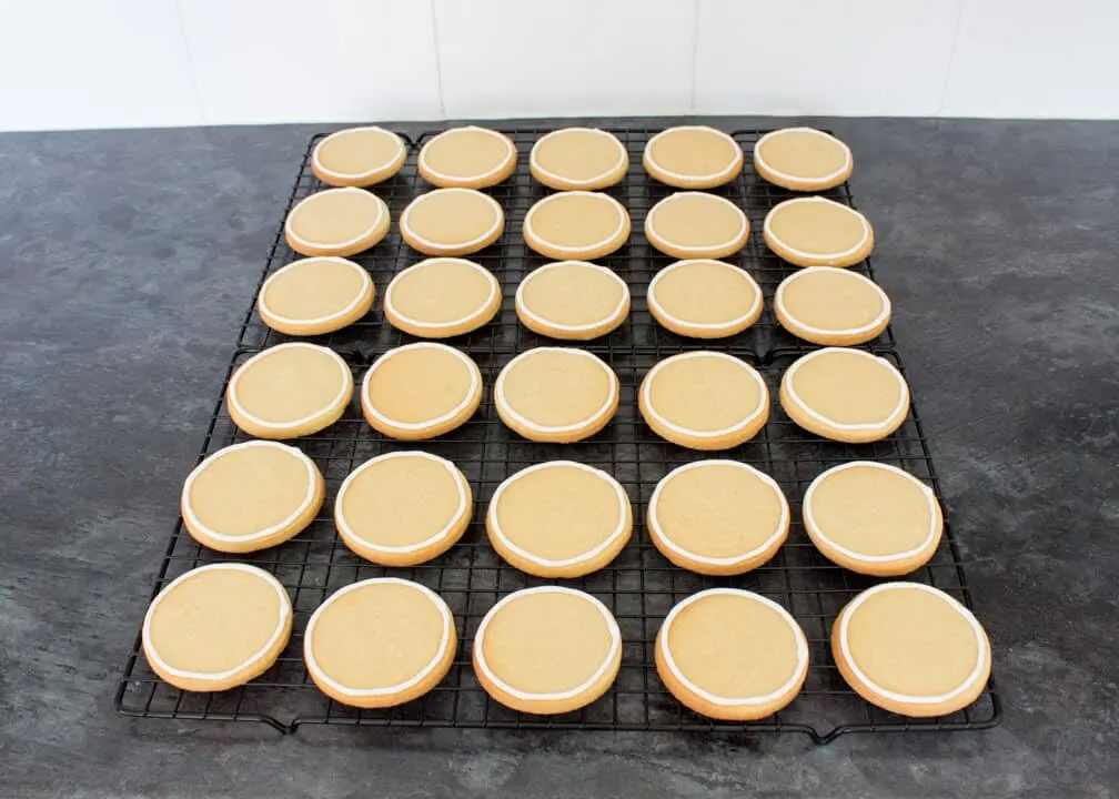 Cookies on cooling racks on a kitchen worktop that have white royal icing piped round the edges.