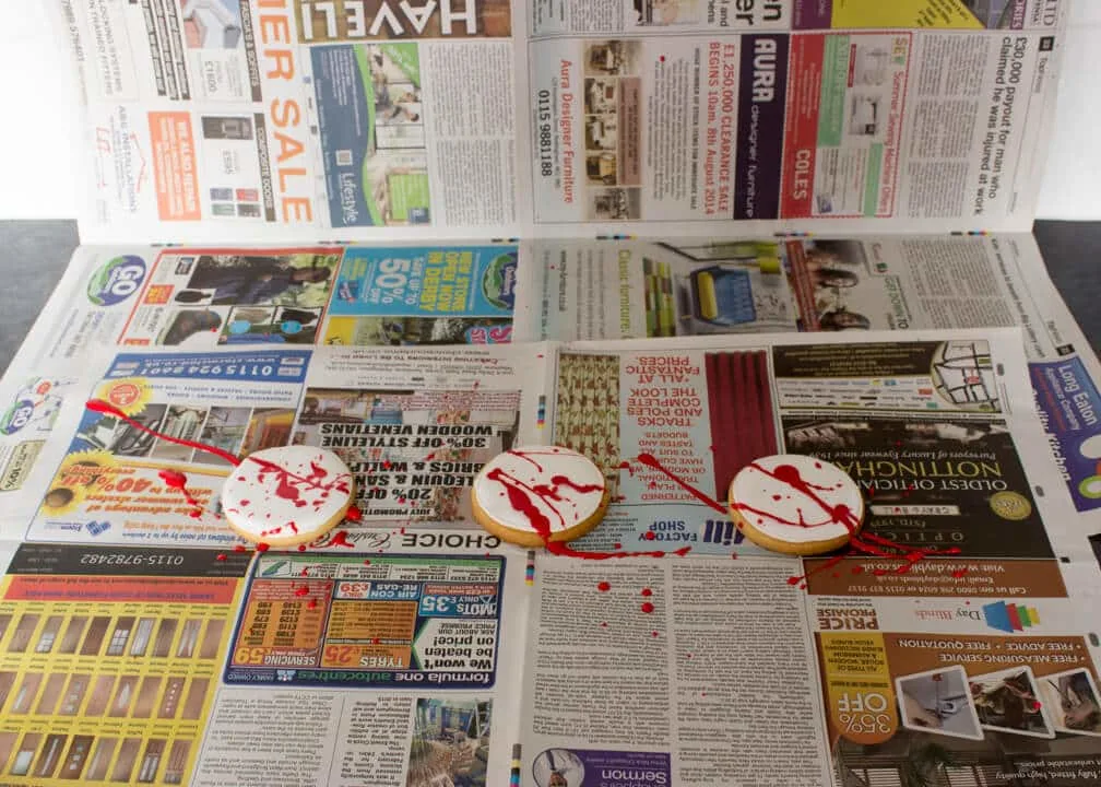 3 cookies on newspaper that have been spattered with blood coloured royal icing.