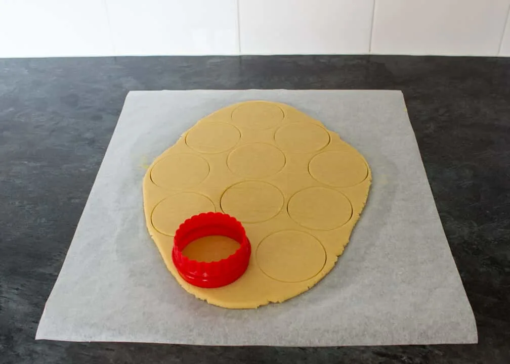 Cookie dough rolled out on a sheet of baking paper with a cookie cutter having cut out lots of circles.
