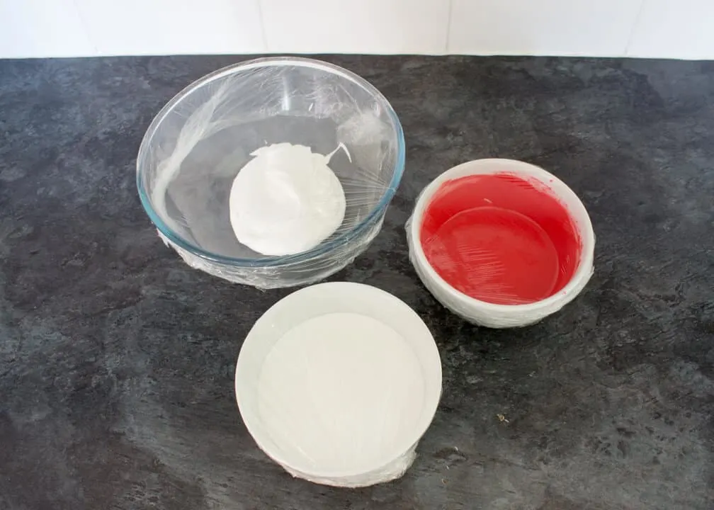 Red flooding royal icing, white flooding royal icing and white stiffer royal icing in bowls covered with cling film on a kitchen worktop.