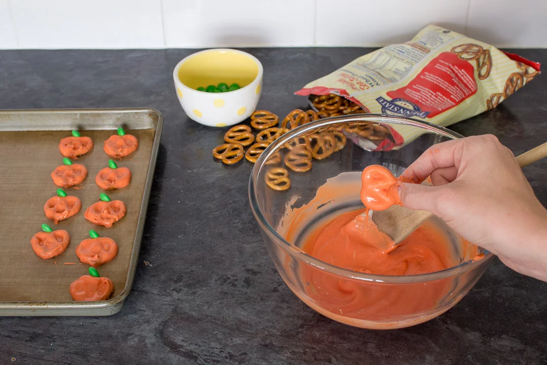 Pretzels being dipped into orange coloured white chocolate then topped with a green M&M and placed onto a baking tray
