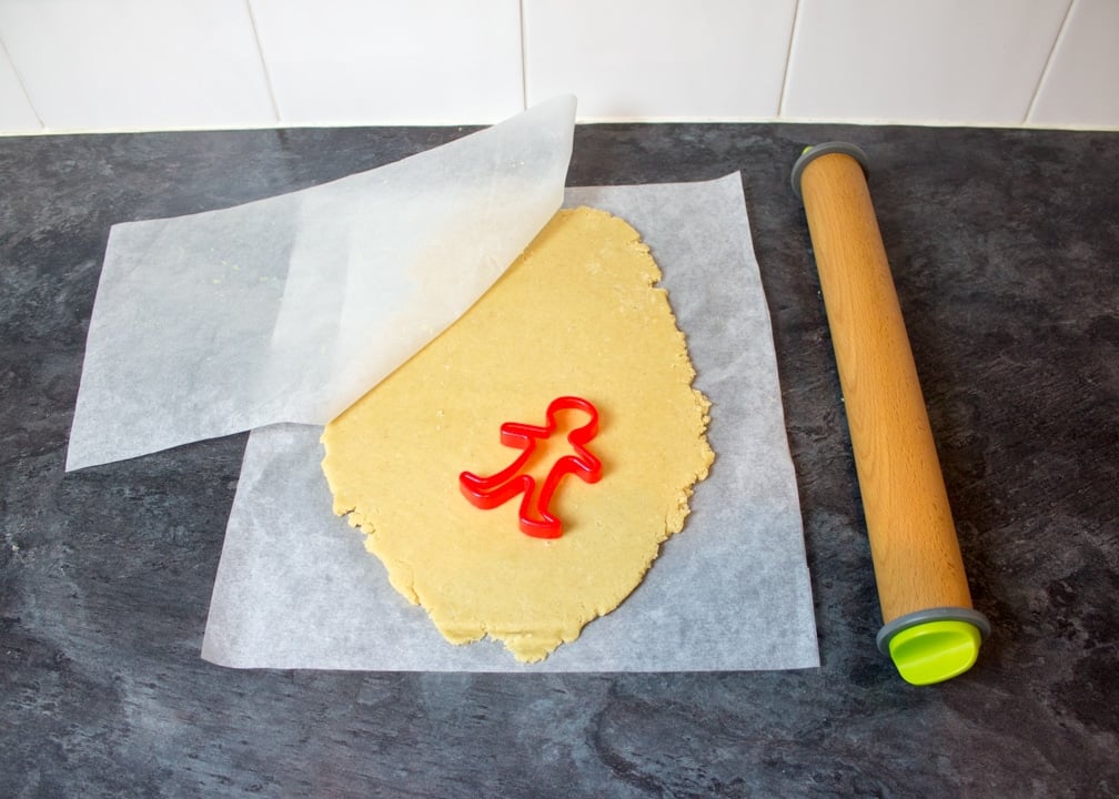 Ginger biscuit dough rolled flat between 2 sheets of baking paper with a gingerbread man cutter on top and a rolling pin on the side. All on a kitchen worktop.