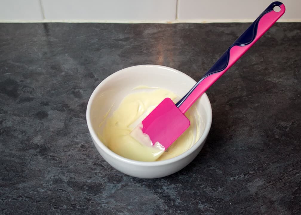 Melted white chocolate in a small bowl with a spatula on a kitchen worktop.