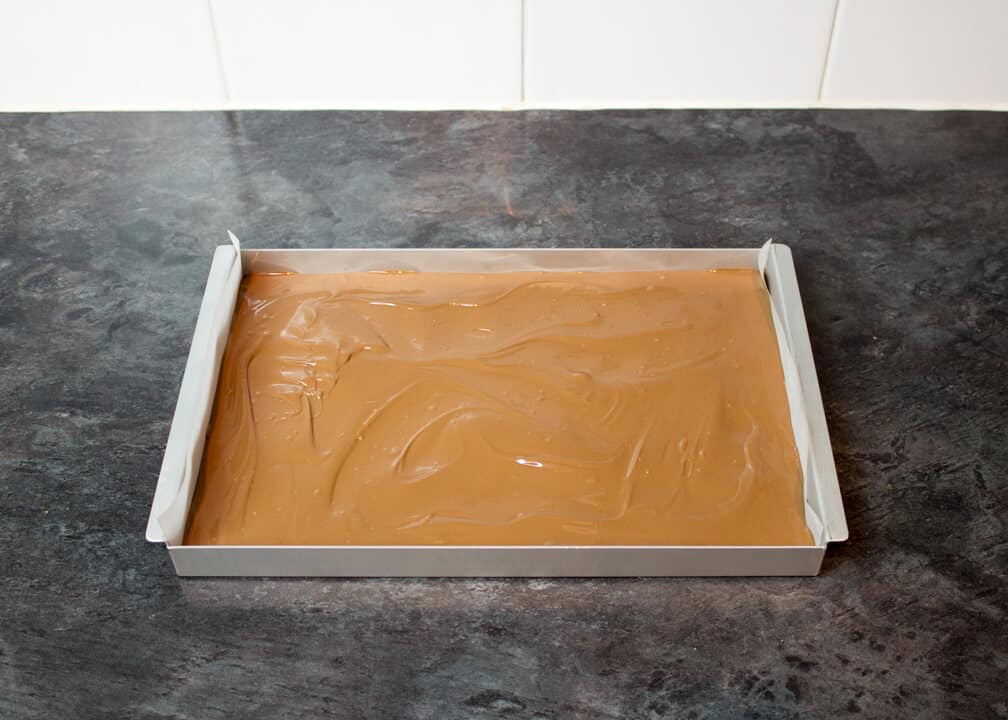 Melted milk chocolate smoothed out in a lined rectangular baking tin on a kitchen worktop.