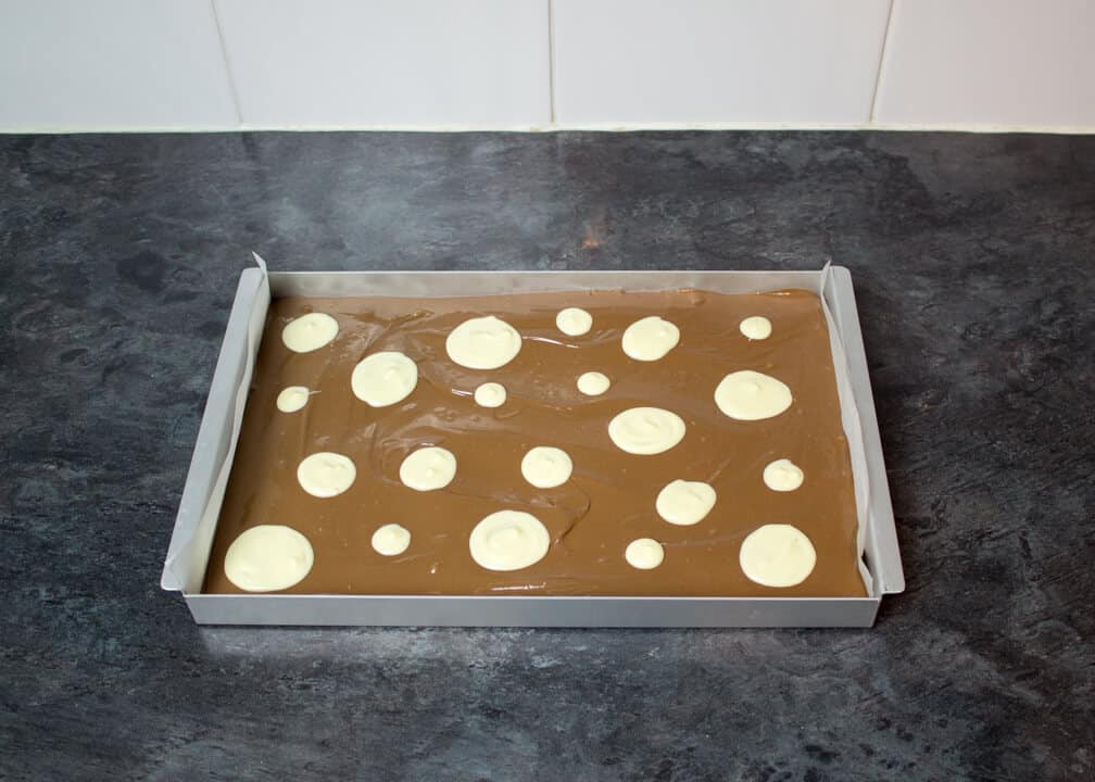 Melted milk chocolate smoothed out in a lined rectangular baking tin with white chocolate circles on top on a kitchen worktop.