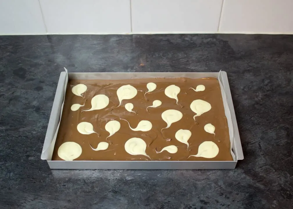 Melted milk chocolate smoothed out in a lined rectangular baking tin with white chocolate ghost shapes on top on a kitchen worktop.