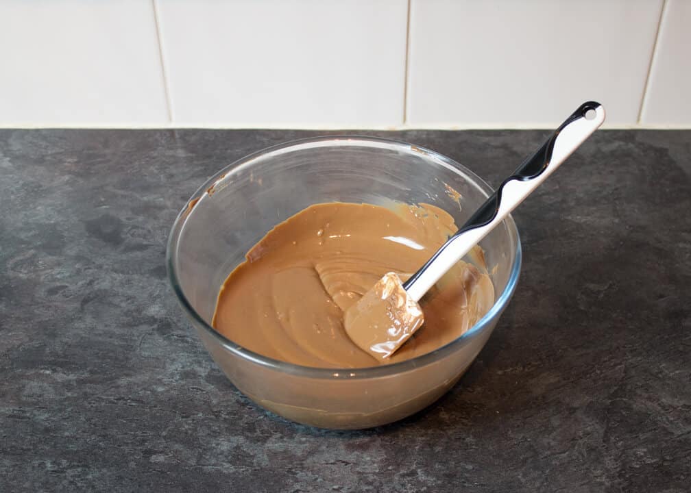 Melted milk chocolate in a glass bowl with a spatula on a kitchen worktop