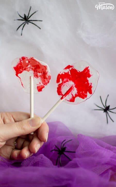 A hand holding 2 bloody Halloween lollipops against a white spider web background behind a base of purple netting. There are 3 black plastic spiders scattered around.