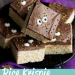 Rice Krispie Halloween treats on a black plate with fake spiders. Set against purple netting on a grey backdrop, there is also a small pumpkin light in the background. A text overlay says " rice krispie halloween treat bars"