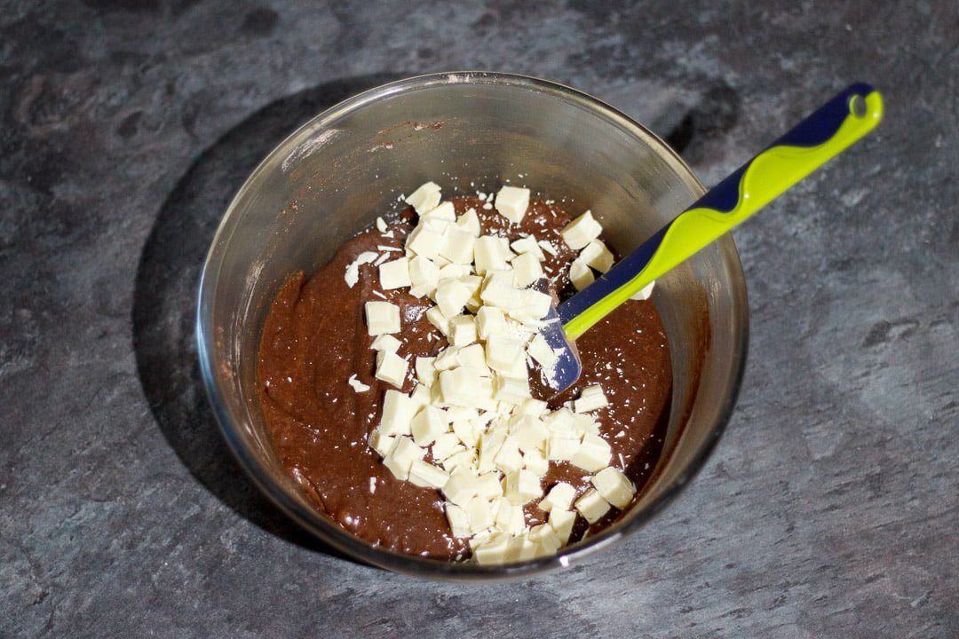 Chocolate brownie batter and white chocolate chunks in a glass bowl with a rubber spatula
