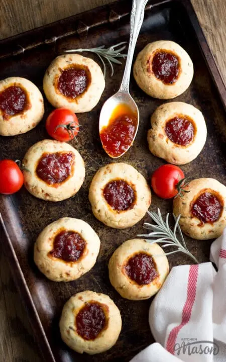 Cheesy Rosemary and Thyme Thumbprint Cookies on a baking tray with a spoon covered in chutney. cherry tomatoes and rosemary in the background.