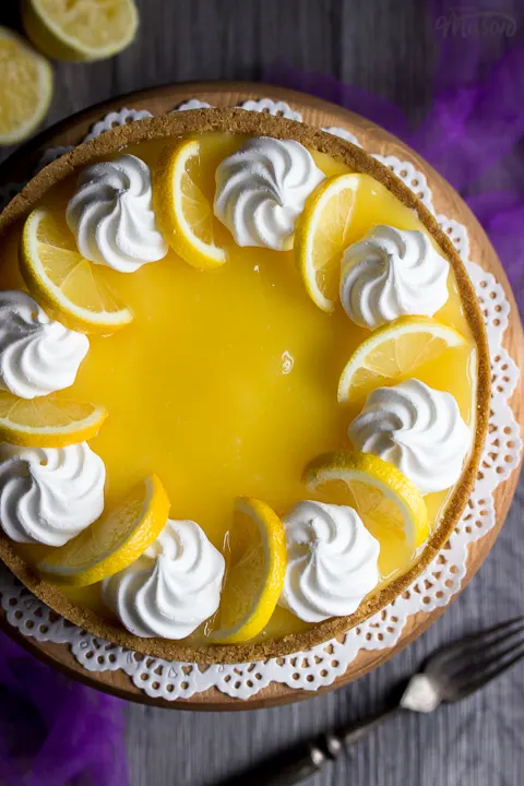 A birds eye view of a whole no bake lemon cheesecake on a cake stand set over purple mesh fabric. Topped with meringue nests and lemon slices.