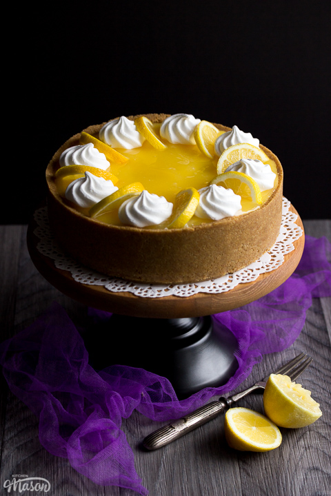 A whole no bake lemon cheesecake on a cake stand set over purple mesh fabric. Topped with meringue nests and lemon slices.