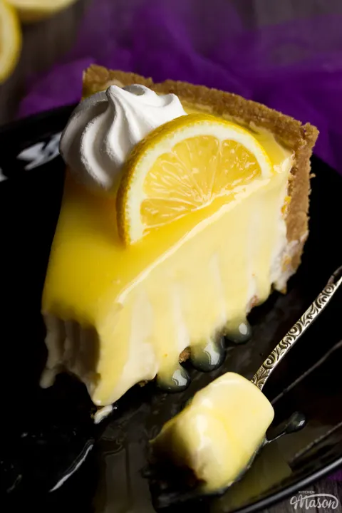A slice of lemon cheesecake on a black plate topped with a meringue nest and lemon slice