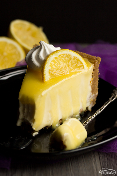A slice of lemon cheesecake on a black plate topped with a meringue nest and lemon slice