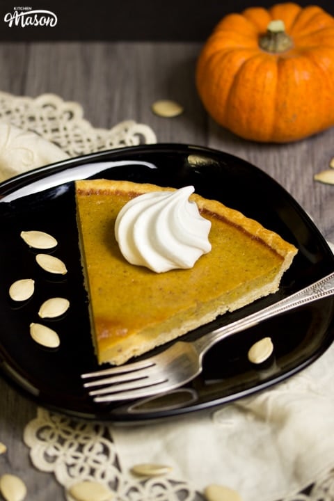 a slice of pumpkin pie with a meringue swirl on top on a black plate with a fork and pumpkin seeds