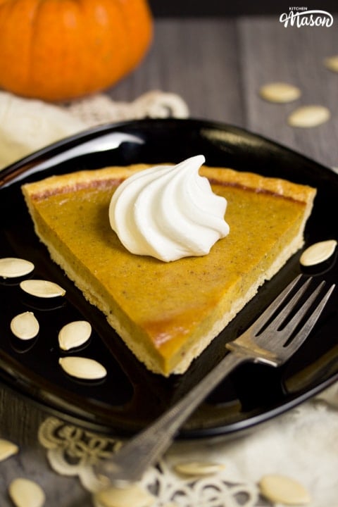 a slice of pumpkin pie with a meringue swirl on top on a black plate with a fork and pumpkin seeds
