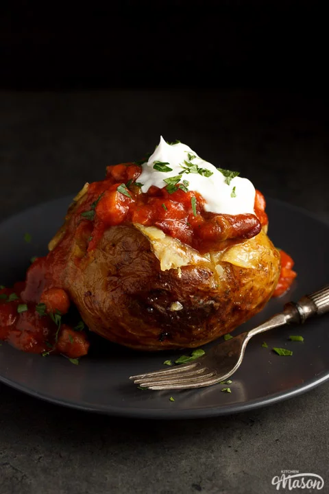 A slow cooker jacket potato on a grey plate topped with vegan chilli and vegan creme fraiche