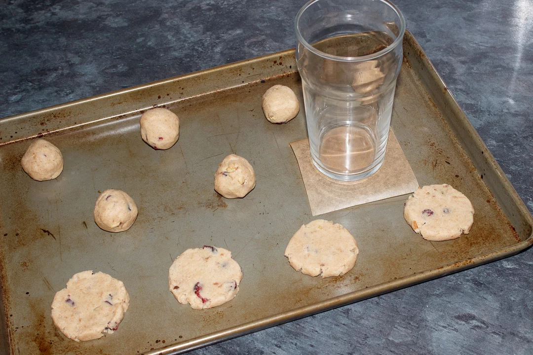 Cranberry Walnut Cinnamon Cookie dough balls on a baking tray being flattened with a glass and a small square of baking paper