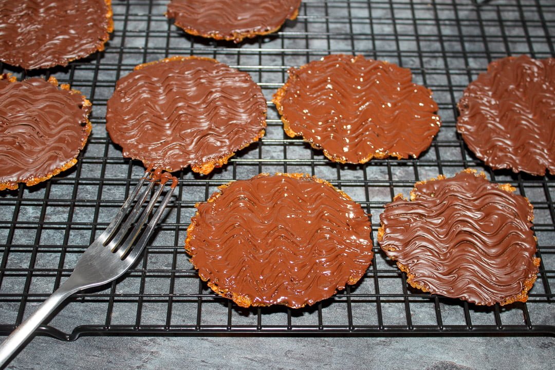 Florentine cookies on a cooling rack topped with melted dark chocolate - using a fork to drag a wavey line pattern into the top.