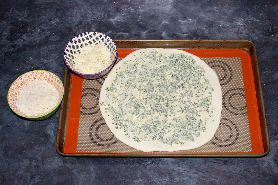 A circle of pizza dough on a large baking tray, covered in herb butter and grated cheese