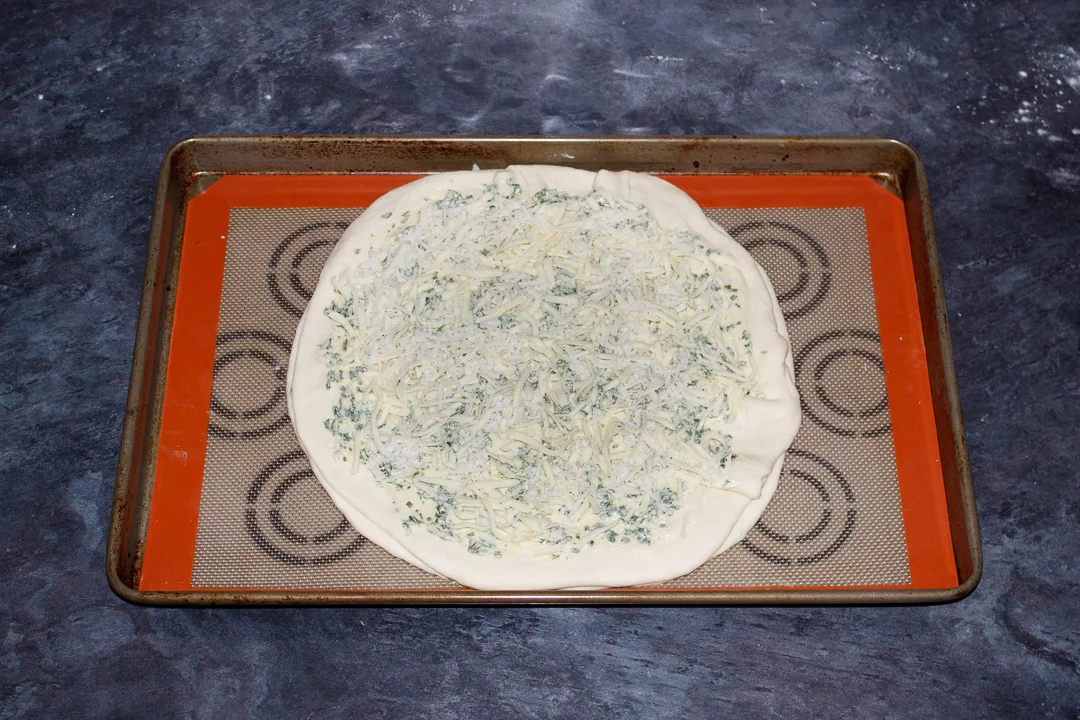 A circle of pizza dough on a large baking tray, covered in herb butter and grated cheese