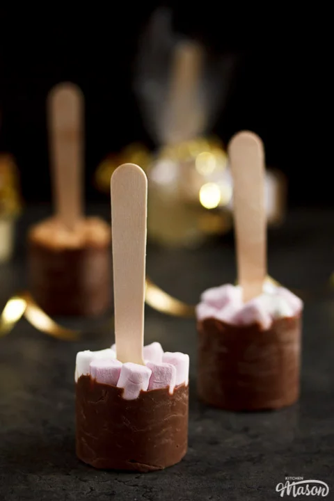 Hot chocolate sticks topped with fudge pieces and marshmallows surrounded by gold curling ribbon