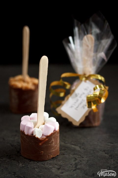 Hot chocolate sticks topped with fudge pieces and marshmallows surrounded by gold curling ribbon