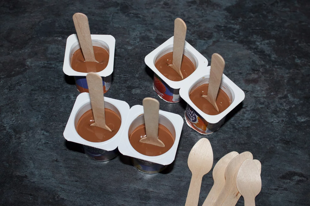 Melted milk chocolate in small yoghurt pots with wooden teaspoons pressed in the middle