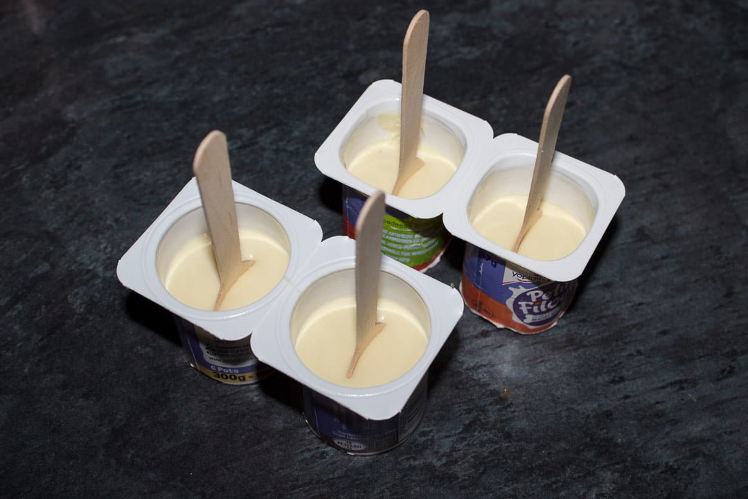 Melted white chocolate in small yoghurt pots with wooden teaspoons pressed in the middle