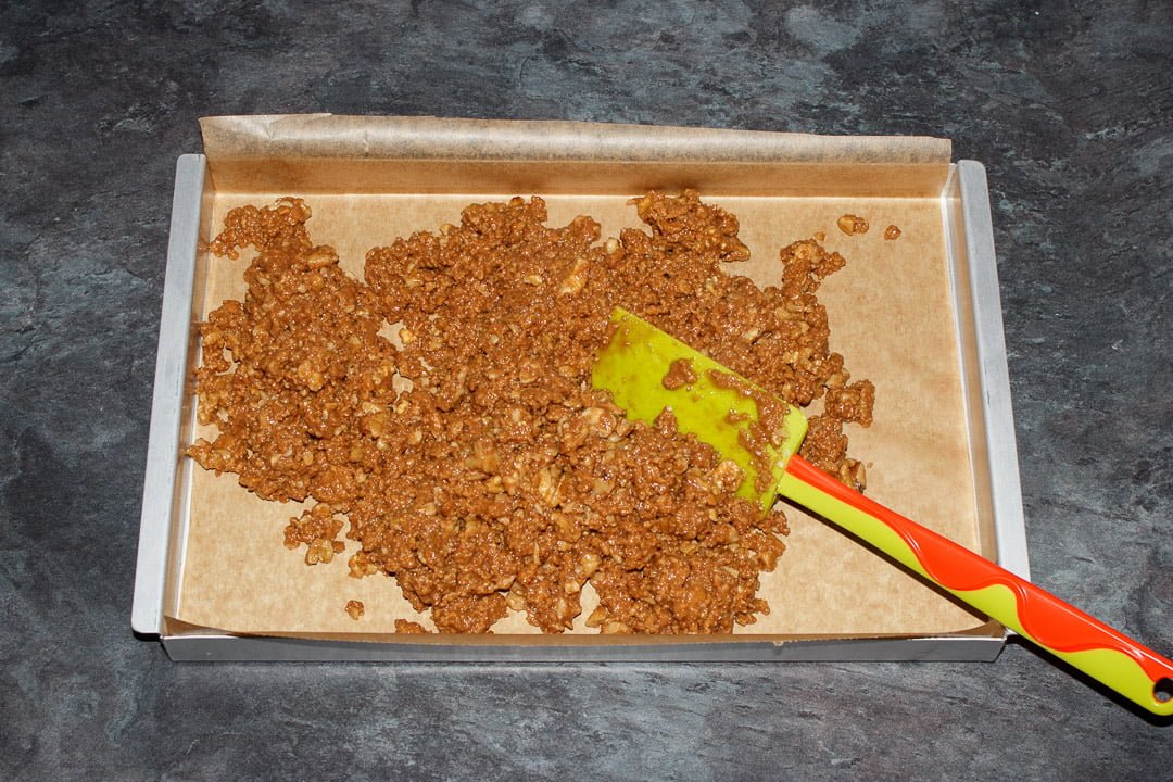 Tiffin recipe mixture being pressed into a rectangular baking tin with a green spatula