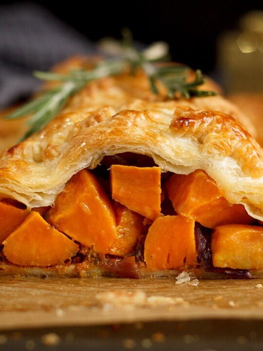 Sweet potato cubes and red onion falling out of a vegetable wellington topped with rosemary sprigs