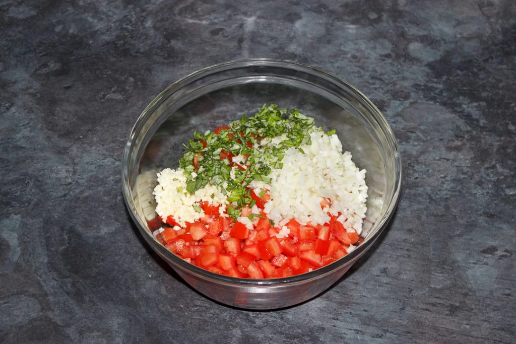 Deseeded chopped tomato in a glass bowl with diced onion, garlic and fresh basil.