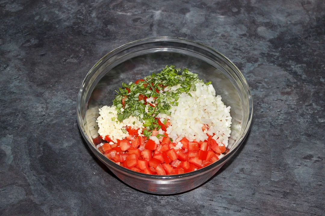 Deseeded chopped tomato in a glass bowl with diced onion, garlic and basil