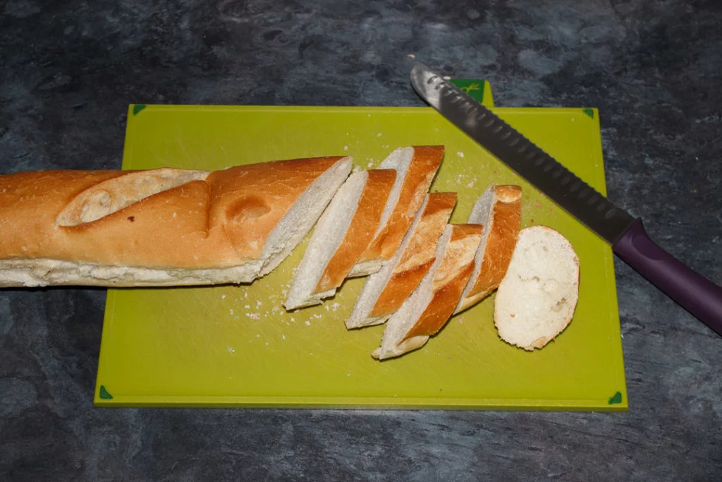 A large baguette being sliced with a bread knife on a green chopping board.
