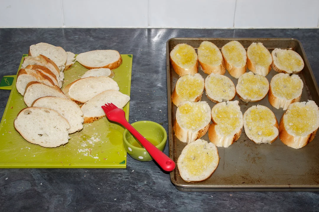 Slices of baguette being brushed on both sides with olive oil then placed onto a baking sheet