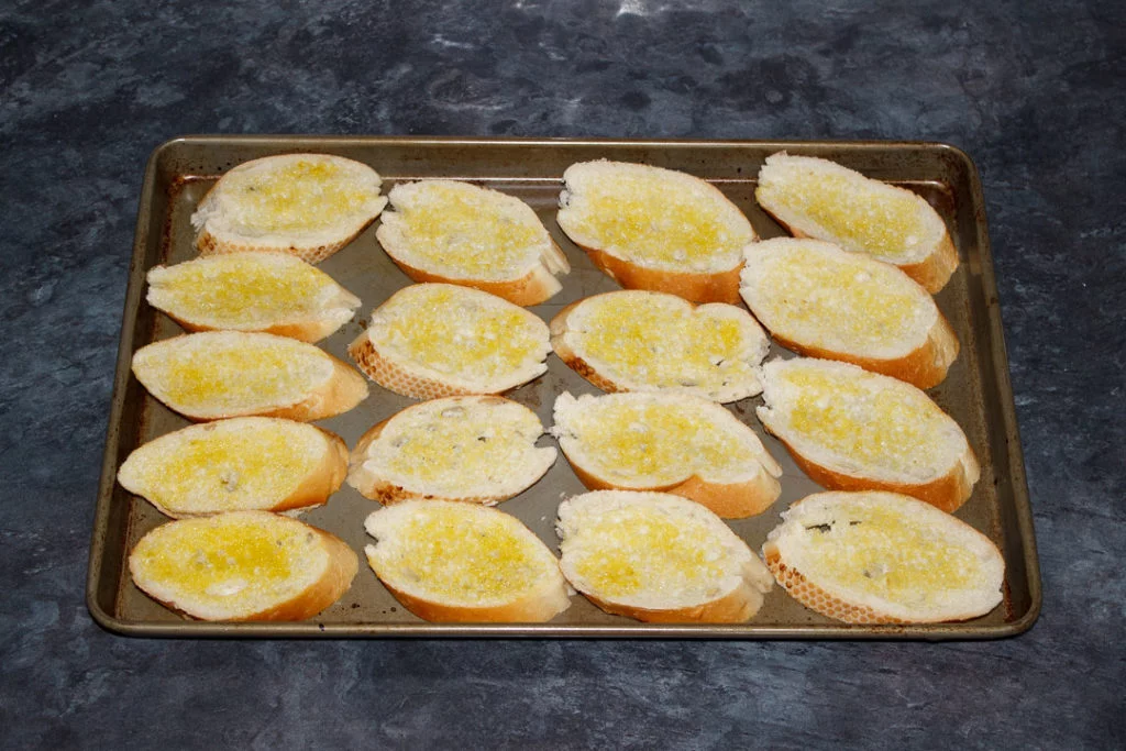 A large baking sheet filled with oiled baguette slices.