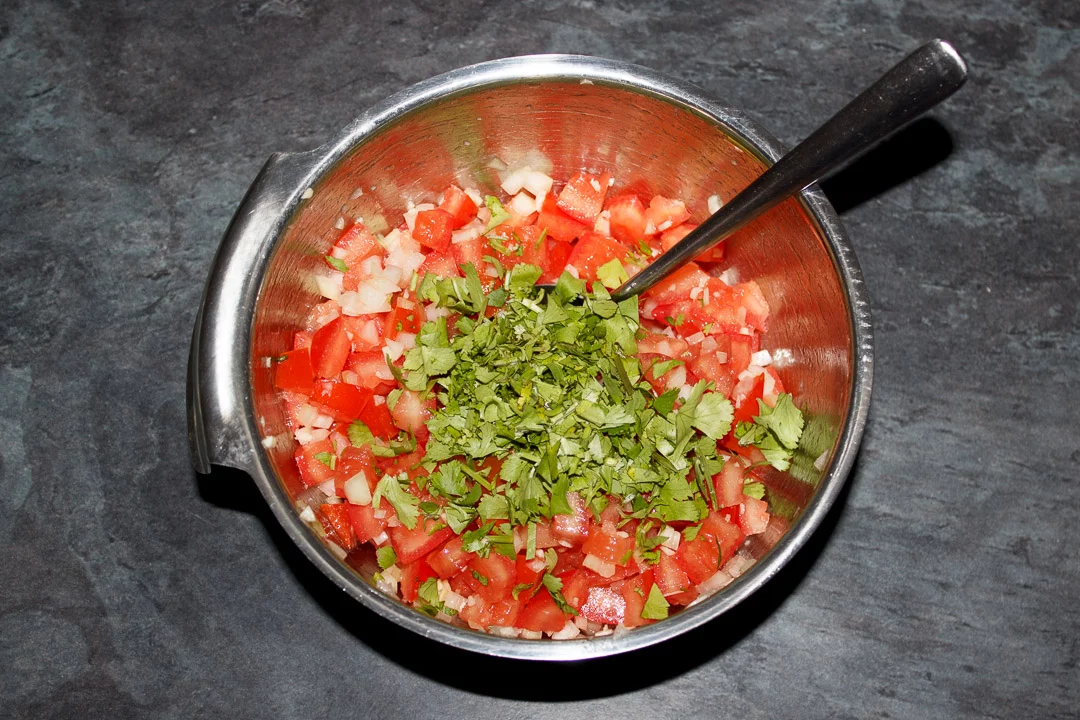 Diced desseded tomato, diced onion and garlic mixed together in a silver bowl, topped with freshly chopped coriander