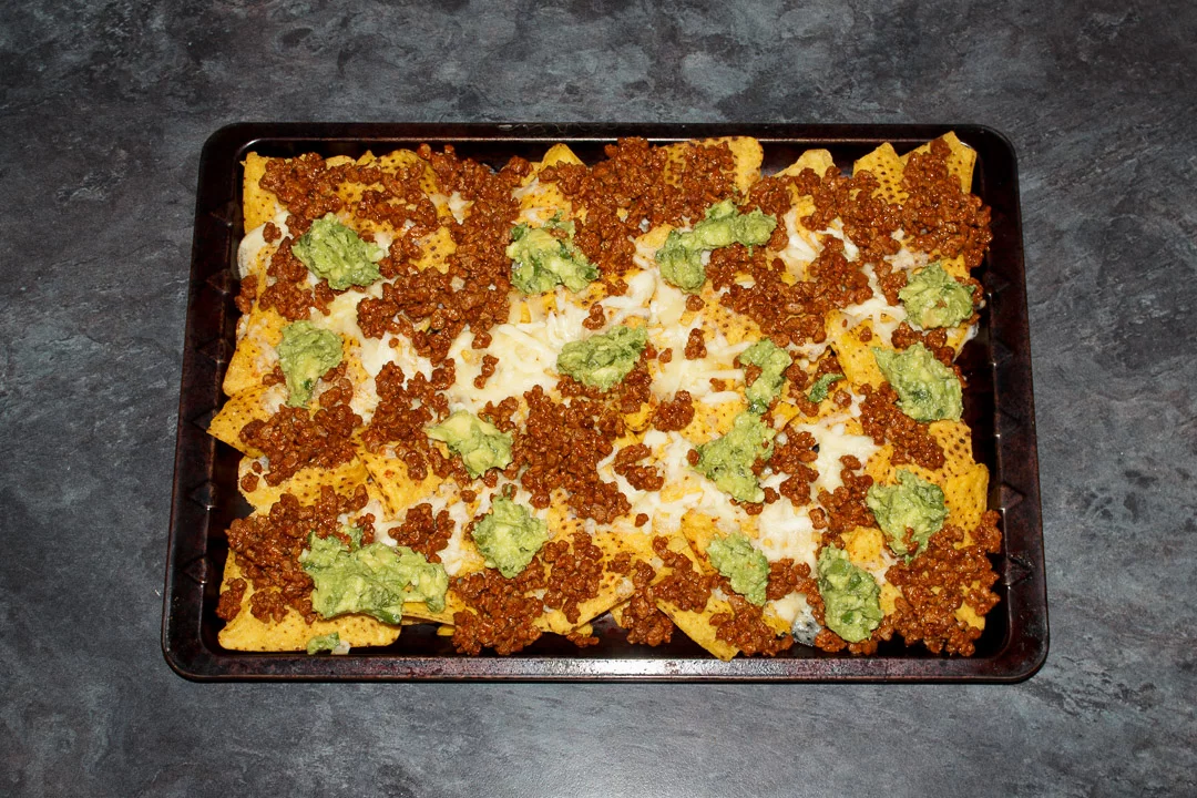 Tortilla chips spread out on a baking tray covered in melted cheese, taco seasoned soya mince and guacamole