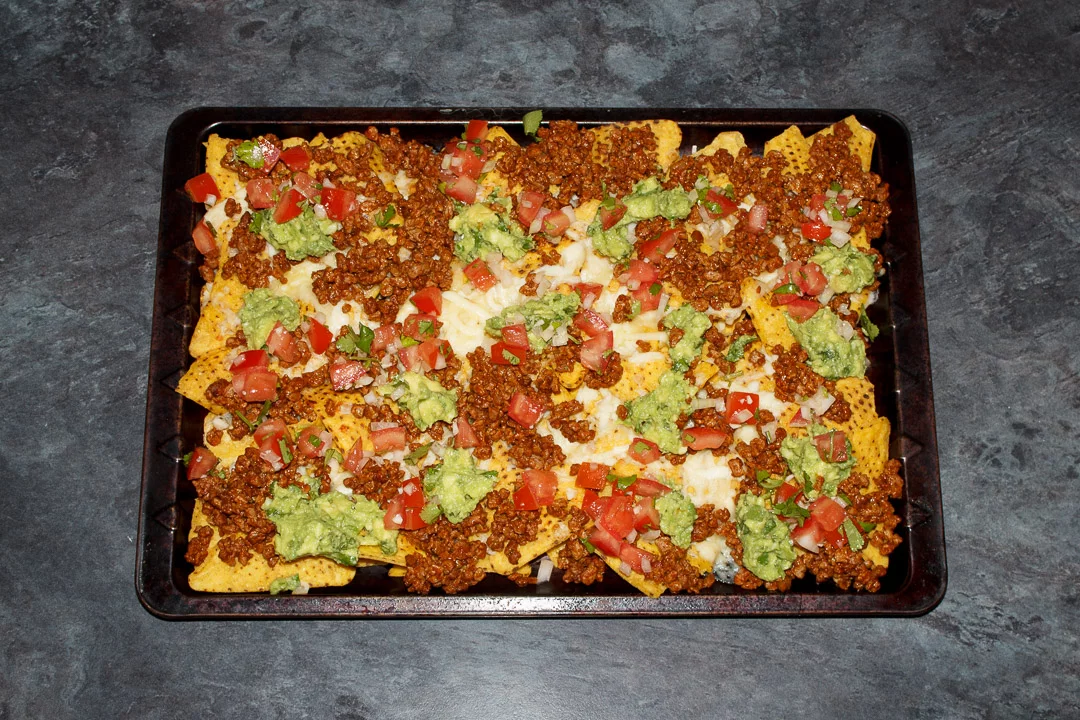 Tortilla chips spread out on a baking tray covered in melted cheese, taco seasoned soya mince, guacamole and tomato salsa