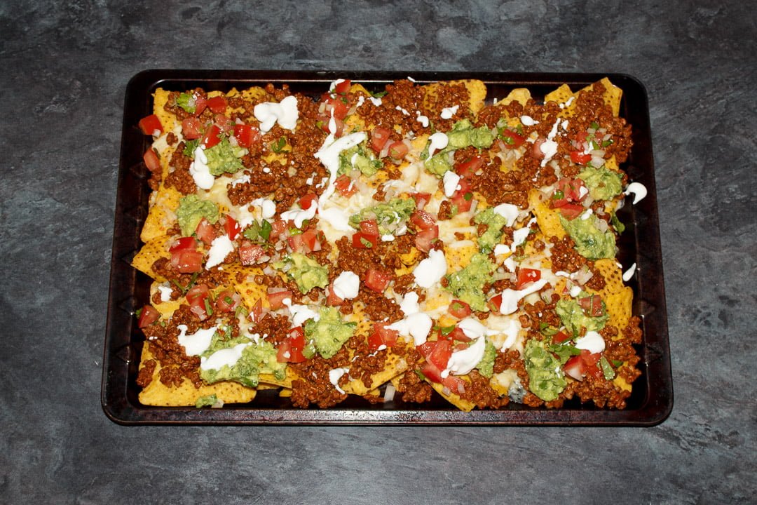 Tortilla chips spread out on a baking tray covered in melted cheese, taco seasoned soya mince, guacamole, tomato salsa and sour cream