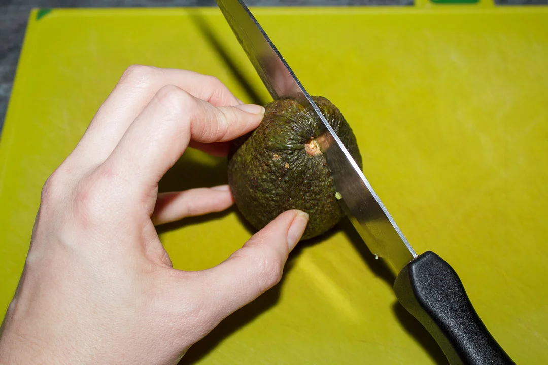 An avocado being sliced in half with a sharp knife on a green chopping board