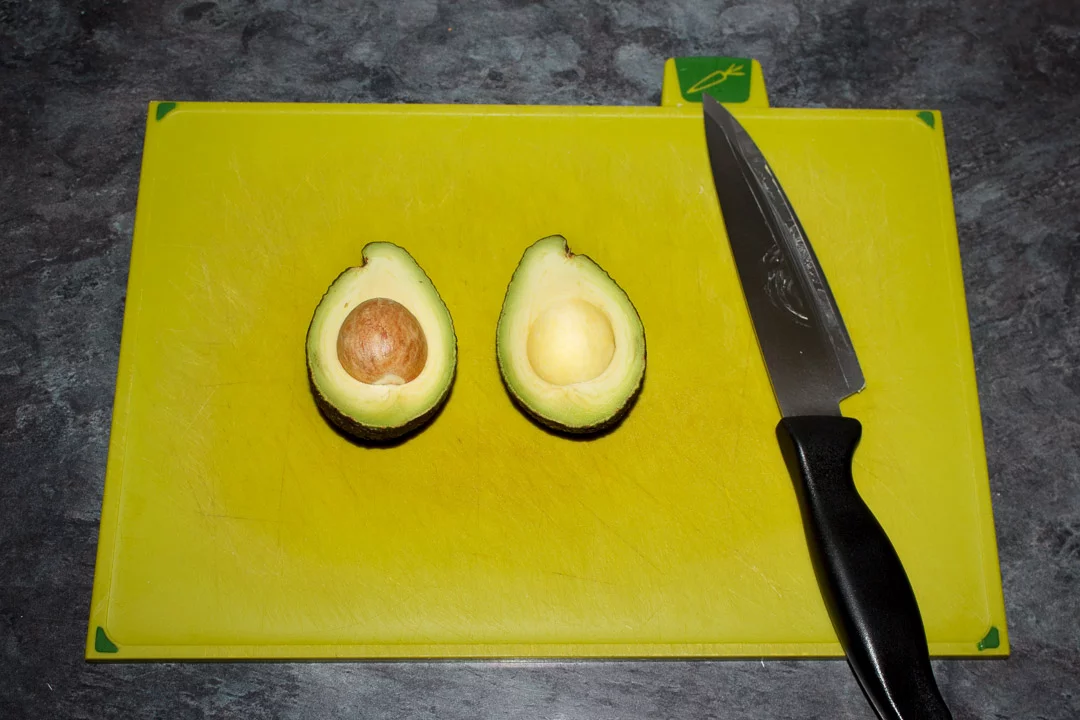 a halved avocado on a chopping board with a sharp knife