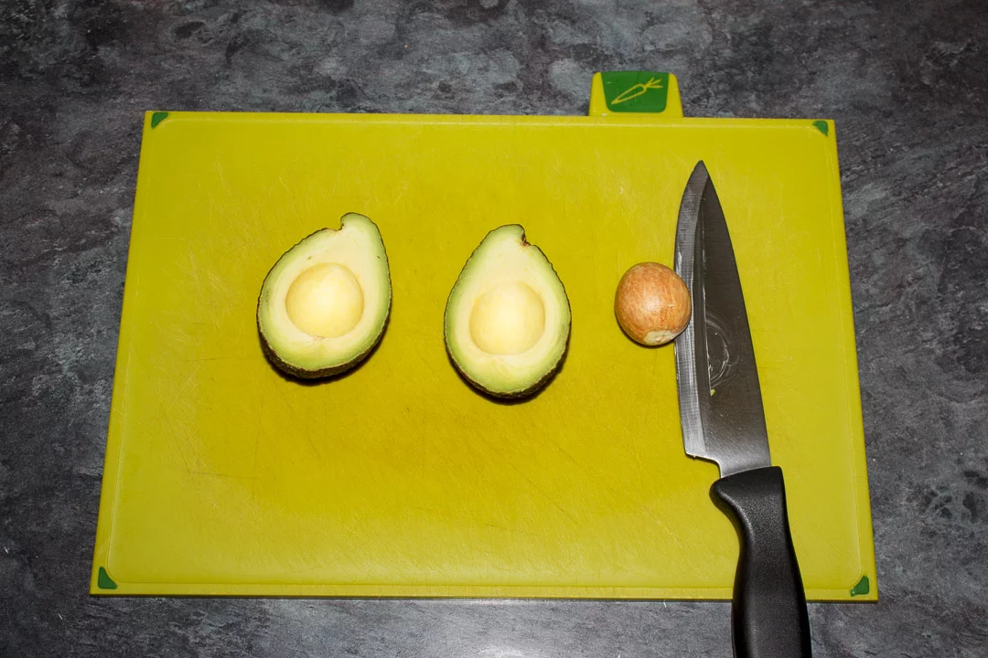 A halved avocado on a green chopping board with a knife and the avocado pip