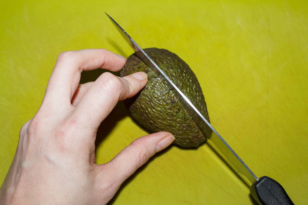 An avocado being sliced in half with a sharp knife on a green chopping board