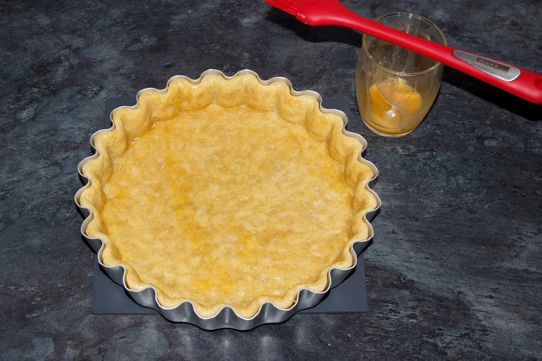 A blind baked shortcrust pastry case in a tart tin with the baking beans removed and beaten egg brushed over it