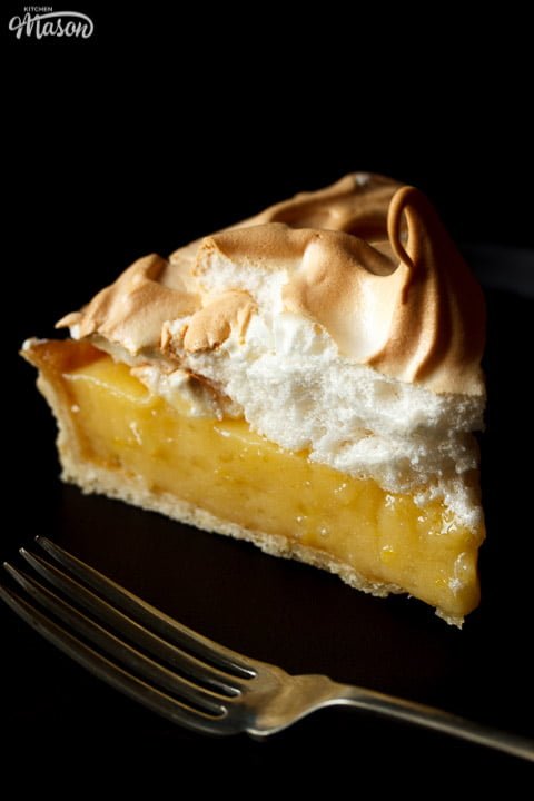 A lemon meringue pie slice on a black plate with a fork