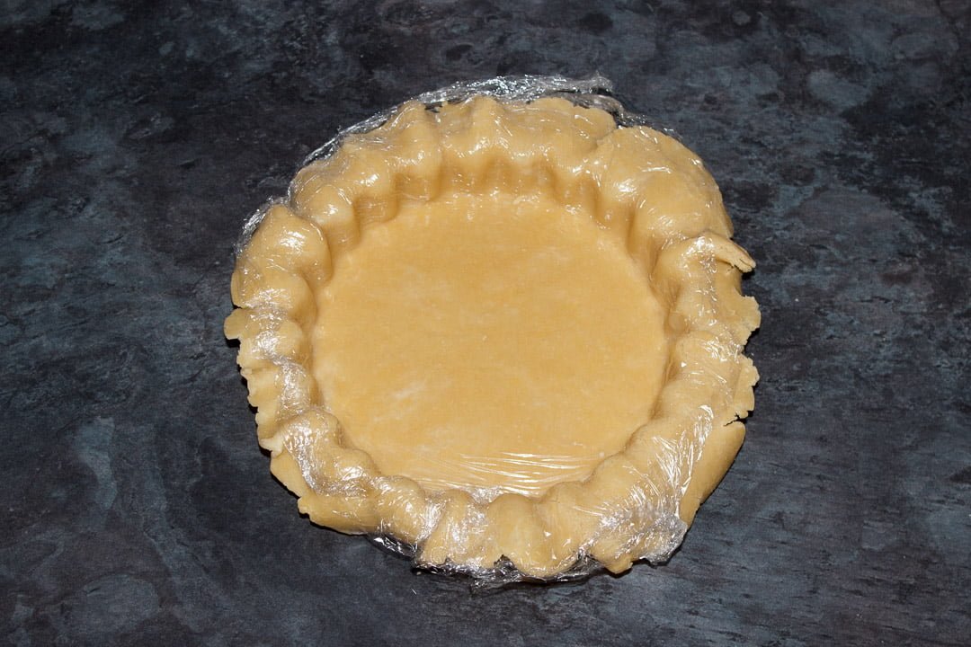 Shortcrust pastry crust being pressed into a tart tin with a piece of cling film